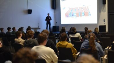 prelegent stojący na scenie z mikrofonem w ręku, prowadzący prezentację przed grupą osób siedzących na widowni. Na ekranie wyświetlana jest wizualizacja budynku w formie rzutu architektonicznego. Publiczność składa się głównie z młodych osób, które uważnie słuchają. Sala jest nowoczesna, z minimalistycznym wystrojem, a scena wyposażona jest w podest i mównicę.