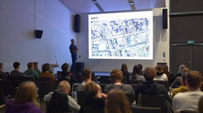 Na zdjęciu przedstawiono prezentację w sali konferencyjnej. Na ekranie wyświetlona jest mapa fragmentu miasta, co wskazuje na omawianie zagadnień urbanistycznych lub architektonicznych. Prowadzący, stojąc z mikrofonem, wyjaśnia treść slajdu. W pierwszym planie widać uczestników prezentacji siedzących na krzesłach i skupionych na wykładzie. Scena oświetlona jest jasnym światłem, tworząc profesjonalną atmosferę spotkania.