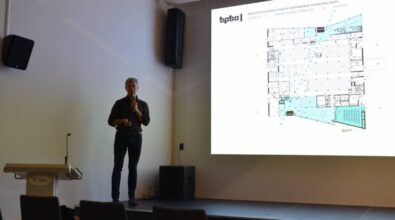 prelegent stojący na scenie z mikrofonem, omawiający plan funkcjonalny parteru budynku widoczny na slajdzie prezentacji. Scena znajduje się w nowoczesnej sali konferencyjnej, a w tle widoczny jest minimalistyczny wystrój wnętrza oraz ekran z wyraźnym rzutem technicznym projektu biblioteki publicznej.
