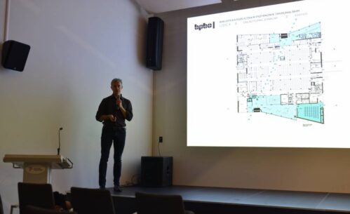 prelegent stojący na scenie z mikrofonem, omawiający plan funkcjonalny parteru budynku widoczny na slajdzie prezentacji. Scena znajduje się w nowoczesnej sali konferencyjnej, a w tle widoczny jest minimalistyczny wystrój wnętrza oraz ekran z wyraźnym rzutem technicznym projektu biblioteki publicznej.