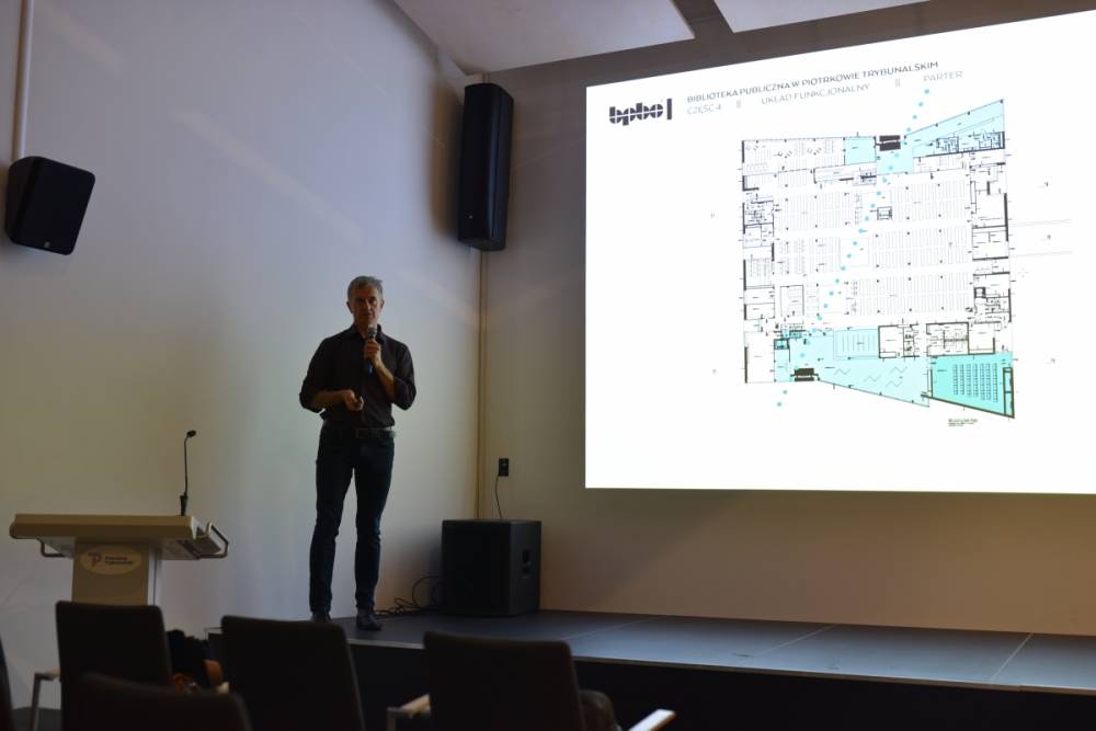 prelegent stojący na scenie z mikrofonem, omawiający plan funkcjonalny parteru budynku widoczny na slajdzie prezentacji. Scena znajduje się w nowoczesnej sali konferencyjnej, a w tle widoczny jest minimalistyczny wystrój wnętrza oraz ekran z wyraźnym rzutem technicznym projektu biblioteki publicznej.