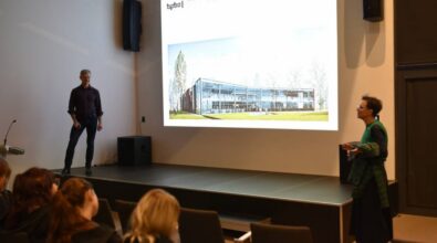 dwie osoby prowadzące prezentację na scenie w sali konferencyjnej. Na ekranie wyświetlony jest wizualny projekt budynku biblioteki publicznej, otoczonego zielenią. Po lewej stronie stoi mężczyzna w ciemnym stroju, a po prawej kobieta w zielono-czarnym ubraniu gestykuluje, prowadząc część prelekcji. W pierwszym planie widoczni są uczestnicy siedzący w rzędach krzeseł.