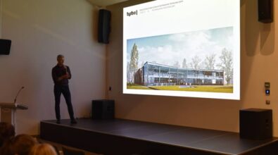 mężczyzna stojący na scenie z mikrofonem w ręku. Po jego prawej stronie na dużym ekranie wyświetlona jest wizualizacja budynku biblioteki publicznej w Piotrkowie Trybunalskim z podpisem: „Rozwiązania materiałowe”. Sala jest przestronna, a na pierwszym planie widoczne są osoby siedzące w rzędach krzeseł.