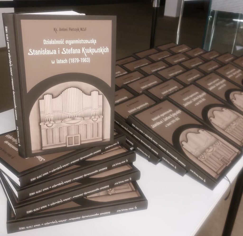 Na pierwszym planie wyeksponowany jest egzemplarz książki o tytule: „Działalność organmistrzowska Stanisława i Stefana Kukukowskich w latach (1879–1963)” autorstwa ks. Antoniego Pietrzyka M.S.F. Okładka jest w stonowanych odcieniach brązu, z grafiką przedstawiającą organy kościelne w centralnej części. W tle widoczne są kolejne egzemplarze książki starannie poukładane w rzędach.