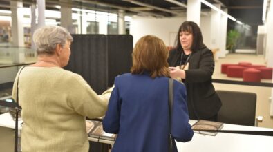 Na zdjęciu widzimy dwie kobiety, które podchodzą do stanowiska obsługi. Stoją tyłem do obiektywu, jedna w jasnym swetrze, a druga w niebieskiej marynarce. Po drugiej stronie stołu znajduje się kobieta ubrana na czarno, podająca jeden z egzemplarzy książki. Na stole leżą książki z widoczną okładką, gdyż scena odbywa się podczas wydarzenia związanego z ich promocją. W tle widać wnętrze nowoczesnej przestrzeni, z czerwonymi pufami oraz jasnym oświetleniem.