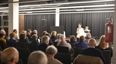 Na zdjęciu widzimy prelekcję, która odbywa się w sali konferencyjnej. Na pierwszym planie widoczni są uczestnicy siedzący na krzesłach, zwróceni twarzami w stronę prelegentki. Kobieta w jasnej bluzce stoi na scenie z mikrofonem w ręku, przemawiając do zgromadzonej publiczności. W tle za nią widać czarną kotarę oraz stolik, na którym znajduje się książka. Scena oświetlona jest jasnym, nowoczesnym oświetleniem, a atmosfera wskazuje na oficjalne wydarzenie, takie jak promocja książki