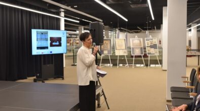 Na zdjęciu widzimy moment promocji książki „Działalność organmistrzowska Stanisława i Stefana Kukukowskich w latach (1879–1963)” autorstwa ks. Antoniego Pietrzyka M.S.F. Kobieta ubrana w elegancką białą bluzkę przemawia, trzymając mikrofon oraz kartki z notatkami. Stoi na tle sceny z czarną kurtyną. Obok znajduje się ekran, na którym wyświetlane są ilustracje związane z tematem wydarzenia. W tle widoczne są plansze wystawy z przedstawieniami organów. W prawym dolnym rogu kadru siedzi uczestniczka wydarzenia z książką w dłoni. Całość odbywa się w przestronnym i nowoczesnym wnętrzu.