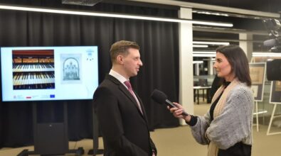 Na zdjęciu widać mężczyznę w eleganckim garniturze z bordowym krawatem, udzielającego wywiadu kobiecie z mikrofonem w ręku. Kobieta ubrana jest w czarny strój i szary sweter, a w tle widoczny jest ekran prezentujący zdjęcia klawiatury organowej oraz szkic organów. W tle można również zauważyć elementy wystawy oraz plansze na sztalugach. Całość ma miejsce podczas promocji książki „Działalność organmistrzowska Stanisława i Stefana Kukukowskich w latach (1879–1963)” w nowoczesnym wnętrzu z czarnymi zasłonami i jasnym oświetleniem.