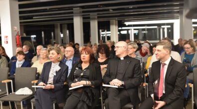 Na zdjęciu widzimy zgromadzoną publiczność podczas wydarzenia kulturalnego. W pierwszym rzędzie siedzi kilka osób w eleganckich strojach: kobieta w granatowym kostiumie z apaszką w kropki, kobieta w czarnej sukni z biżuterią, duchowny w sutannie oraz mężczyzna w garniturze i czerwonym krawacie. Każda z tych osób trzyma w rękach książkę. W tle widać kolejne rzędy uczestników, siedzących w nowoczesnej sali z dużymi oknami i oświetleniem sufitowym