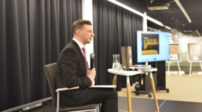 Na zdjęciu widzimy mężczyznę w eleganckim garniturze z czerwonym krawatem, siedzącego na krześle na scenie podczas wystąpienia. Trzyma mikrofon i patrzy w stronę publiczności, uśmiechając się. Na stoliku obok niego znajdują się książki, szklany dzbanek z wodą oraz szklanka. W tle widoczny jest ekran prezentacyjny z wyświetloną grafiką oraz tablice z wystawą. Kurtyny i oświetlenie sceniczne podkreślają formalny charakter wydarzenia.