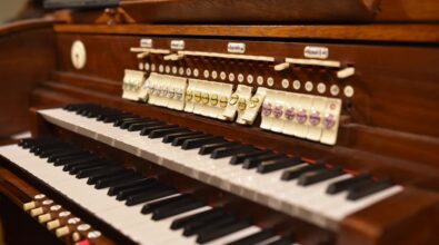 Zbliżenie na klawiaturę oraz rejestry organów piszczałkowych. Widać eleganckie, drewniane wykończenie instrumentu oraz dwa rzędy klawiszy w kolorach czerni i bieli. Powyżej znajdują się kolorowe guziki rejestrów z opisami, które służą do zmiany brzmienia organów. Na górze widoczny jest fragment wyświetlacza z nutami, co wskazuje na wykorzystanie nowoczesnej technologii w tradycyjnym instrumencie.