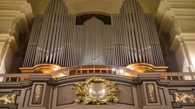 Widok na główny prospekt organowy w kościele. W centralnej części znajdują się rzędy srebrzystych piszczałek o różnej wysokości, tworzące symetryczny i majestatyczny układ. Prospekt jest ozdobiony złotymi detalami oraz dekoracjami rzeźbiarskimi w stylu barokowym. Poniżej widoczny jest ozdobny herb otoczony bogatymi złoceniami, a całość harmonijnie wkomponowana w architekturę wnętrza.