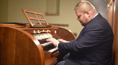 Organista w garniturze gra na organach o klasycznym drewnianym wykończeniu. Instrument posiada kilka klawiatur oraz rzędy przycisków i rejestrów. W tle widoczny jest delikatnie rozmyty fragment wnętrza kościoła, tworząc spokojną i skupioną atmosferę.