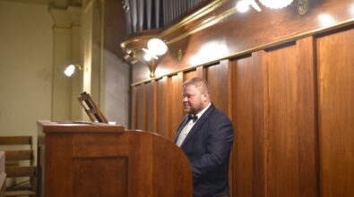 Organista w eleganckim garniturze gra na organach, znajdujących się w klasycznym wnętrzu kościoła. Instrument jest obudowany drewnem, a nad nim widoczna jest część piszczałek. Światło lamp oświetla miejsce gry, podkreślając szczegóły wykończenia. W tle widać fragment drewnianej i złoconej dekoracji.