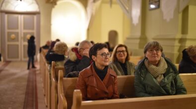 Grupa osób siedząca w drewnianych ławkach w kościele. Na pierwszym planie widoczne dwie kobiety w okularach, jedna w skórzanej kurtce, druga w zielonej kurtce z szalem. W tle pozostała część zgromadzonych, wnętrze oświetlone ciepłym światłem, z ozdobnymi drzwiami i dekoracjami widocznymi w oddali.