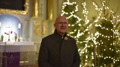 Mężczyzna w okularach, ubrany w ciemny płaszcz, stoi przy mikrofonie w kościele. W tle widoczne ozdobione światełkami choinki oraz ołtarz z krzyżem, kwiatami i złotymi zdobieniami. Jasne światło dodaje uroczystej atmosfery.