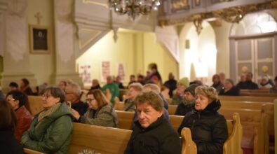 Wnętrze kościoła, wypełnione ludźmi siedzącymi na drewnianych ławkach. Na pierwszym planie widać ludzi w ciepłych kurtkach i szalikach, skupionych na wydarzeniu. W tle dostrzec można elementy dekoracyjne kościoła, takie jak żyrandol, ozdobne filary oraz drzwi. Atmosfera jest spokojna i uroczysta.