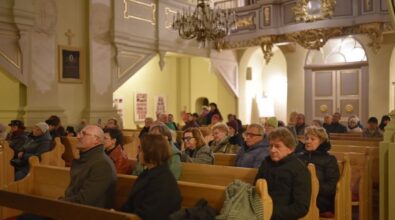 Wnętrze kościoła, wypełnione ludźmi siedzącymi na drewnianych ławkach. Na pierwszym planie widać ludzi w ciepłych kurtkach i szalikach, skupionych na wydarzeniu. W tle dostrzec można elementy dekoracyjne kościoła, takie jak żyrandol, ozdobne filary oraz drzwi. Atmosfera jest spokojna i uroczysta.