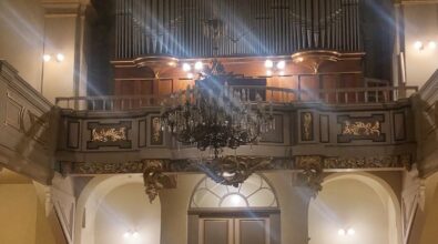 Wnętrze kościoła, widok na tylną część nawy głównej. W centralnej części znajdują się duże organy kościelne oraz balkon z ozdobnymi złoceniami. Nad balkonem widoczny jest żyrandol, a poniżej drzwi prowadzące do zakrystii. Po bokach siedzą wierni w drewnianych ławkach, uczestniczący w wydarzeniu. Jasne oświetlenie podkreśla szczegóły architektury.