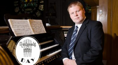 Mężczyzna w garniturze i krawacie, siedzący przy organach kościelnych. W tle znajdują się kolorowe witraże, które tworzą nastrojowe oświetlenie wnętrza kościoła. Na stojaku organowym widnieje otwarta partytura muzyczna. W lewym dolnym rogu zdjęcia znajduje się logo z napisem „Krukowski Day” oraz stylizowanym motywem piszczałek organowych.