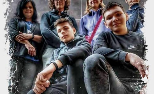 Na zdjęciu widoczna jest grupa sześciu osób pozujących przed szarym, gładkim tłem. Osoby stoją w różnych pozach, na przodzie dwóch młodych mężczyzn siedzi na ugiętych nogach, podczas gdy pozostali stoją za nimi. Zdjęcie ma artystyczną ramkę w stylu rozpryskującej się farby, która dodaje dynamicznego efektu. Grupa wygląda na zgraną, a ich różnorodne wyrazy twarzy wskazują na przyjazną atmosferę. Ubrania osób mają głównie ciemne kolory, co kontrastuje z jasną ramką zdjęcia.