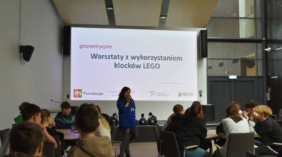 duża aula, na pierwszym planie stoliki wokół których siedzi młodzież, głębiej prowadząca objaśnia coś przez mikrofon. W tle duża ściana z wyświetloną na niej prezentacją