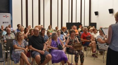 Grupa ludzi siedzi w jasnej sali konferencyjnej, uważnie słuchając osoby, która stoi z przodu z mikrofonem i notatnikiem na klipbordzie. Uczestnicy, głównie dorośli i osoby starsze, wydają się być zaangażowani i zainteresowani prezentacją. W tle widać logo 