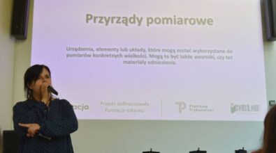 Osoba prowadząca warsztaty stoi na tle ekranu projekcyjnego. Slajd dotyczy przyrządów pomiarowych, na zdjęciu widoczne logotypy Piotrkowa Trybunalskiego, Biblioteki i Fundacji mBanku.