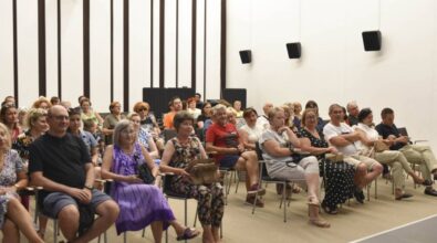 Grupa ludzi siedzi w jasnej sali konferencyjnej, skierowani twarzami w stronę sceny lub ekranu, który znajduje się poza kadrem. Uczestnicy, głównie dorośli i osoby starsze, są skupieni na tym, co się dzieje przed nimi. W sali panuje atmosfera skupienia, a na twarzach niektórych osób widać zainteresowanie lub uśmiechy.
