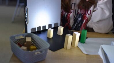 eksperyment edukacyjny z użyciem światła i cieni. Przedstawione są drewniane klocki ustawione na czarnej podstawce oraz ekran z papierem, na którym wyraźnie widać rzucane przez klocki cienie. Obok znajduje się koszyk z dodatkowymi klockami. W tle widoczna jest osoba, uczestnicząca w zajęciach.