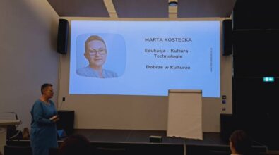 Kobieta w krótkich włosach stoi przed sceną i przemawia, w tle widać ekran projekcyjny oraz tablicę flipchart do szkoleń.