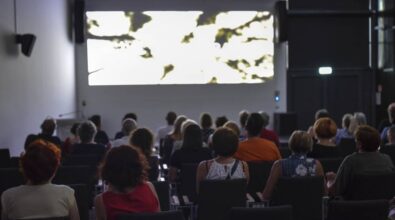 Grupa ludzi siedzi w ciemnej sali kinowej, oglądając film na dużym ekranie. Obraz na ekranie jest jasny i abstrakcyjny, z rozmytymi czarno-białymi kształtami, co sugeruje, że film może mieć artystyczny lub eksperymentalny charakter. Publiczność jest skupiona i w ciszy ogląda projekcję