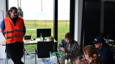 Grupa ludzi znajduje się w pomieszczeniu z dużymi oknami, przez które widać trawiasty teren. Jedna osoba stoi w pomarańczowej kamizelce odblaskowej, pozostałe osoby siedzą przy stole, pracując nad sondą.