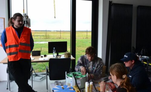 Grupa ludzi znajduje się w pomieszczeniu z dużymi oknami, przez które widać trawiasty teren. Jedna osoba stoi w pomarańczowej kamizelce odblaskowej, pozostałe osoby siedzą przy stole, pracując nad sondą.