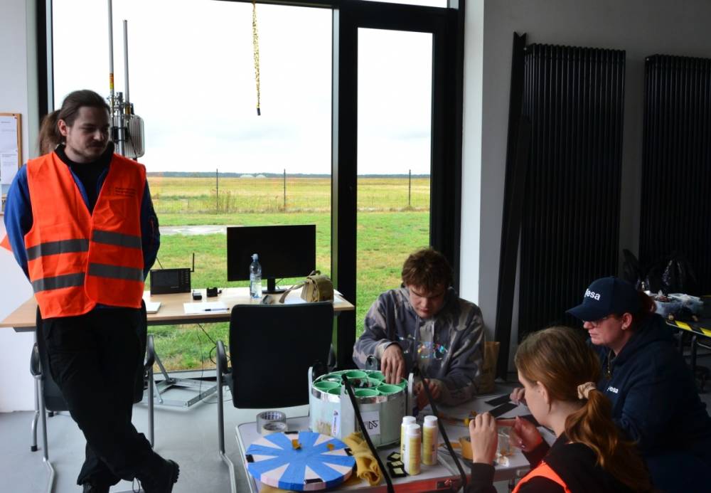 Grupa ludzi znajduje się w pomieszczeniu z dużymi oknami, przez które widać trawiasty teren. Jedna osoba stoi w pomarańczowej kamizelce odblaskowej, pozostałe osoby siedzą przy stole, pracując nad sondą.