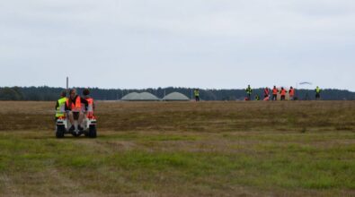 Na pierwszym planie znajduje się mały pojazd z osobami, które mają na sobie kamizelki odblaskowe. W tle widać grupę osób również w kamizelkach odblaskowych na otwartym terenie, przygotowujących się do wypuszczenia dużego balonu meteorologicznego.