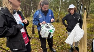 Na zdjęciu znajdują się trzy osoby stojące w lesie. Jedna osoba trzyma urządzenie, które wydaje się być naukowym sprzętem. Inna osoba trzyma biały materiał przypominający spadochron.