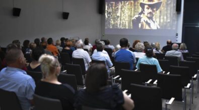 Grupa ludzi siedzi w ciemnej sali kinowej, oglądając projekcję filmu. Na ekranie widoczny jest mężczyzna w kapeluszu kowbojskim, który celuje z pistoletu w stronę kamery, co sugeruje scenę z filmu westernowego. Publiczność, składająca się głównie z dorosłych i starszych osób, jest skupiona na oglądaniu filmu