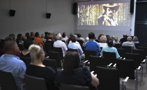 Grupa ludzi siedzi w ciemnej sali kinowej, oglądając projekcję filmu. Na ekranie widoczny jest mężczyzna w kapeluszu kowbojskim, który celuje z pistoletu w stronę kamery, co sugeruje scenę z filmu westernowego. Publiczność, składająca się głównie z dorosłych i starszych osób, jest skupiona na oglądaniu filmu