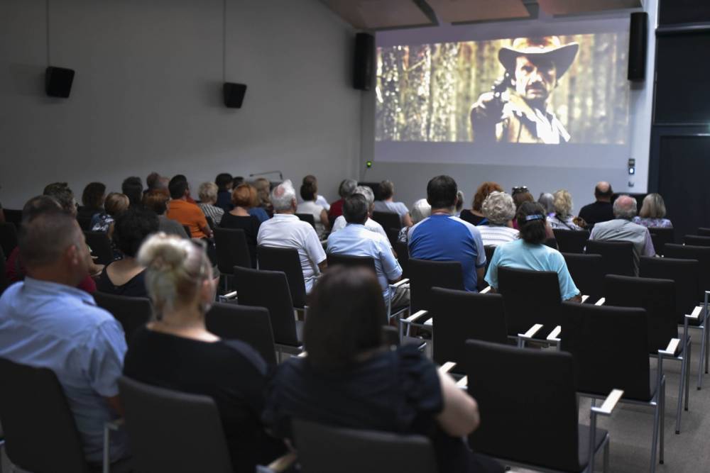 Grupa ludzi siedzi w ciemnej sali kinowej, oglądając projekcję filmu. Na ekranie widoczny jest mężczyzna w kapeluszu kowbojskim, który celuje z pistoletu w stronę kamery, co sugeruje scenę z filmu westernowego. Publiczność, składająca się głównie z dorosłych i starszych osób, jest skupiona na oglądaniu filmu