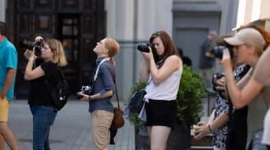 Grupa kobiet stojących na ulicy, każda z nich trzyma aparat fotograficzny i koncentruje się na robieniu zdjęć. Kobiety są różnie ubrane, niektóre mają na sobie letnie stroje, a inne bardziej casualowe. W tle widać fragmenty budynków oraz nieco rozmyte sylwetki przechodniów. Każda z fotografek wydaje się być skupiona na swoim celu, różne kąty ustawienia aparatów sugerują, że każda z nich fotografuje coś innego.