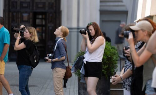 Grupa kobiet stojących na ulicy, każda z nich trzyma aparat fotograficzny i koncentruje się na robieniu zdjęć. Kobiety są różnie ubrane, niektóre mają na sobie letnie stroje, a inne bardziej casualowe. W tle widać fragmenty budynków oraz nieco rozmyte sylwetki przechodniów. Każda z fotografek wydaje się być skupiona na swoim celu, różne kąty ustawienia aparatów sugerują, że każda z nich fotografuje coś innego.