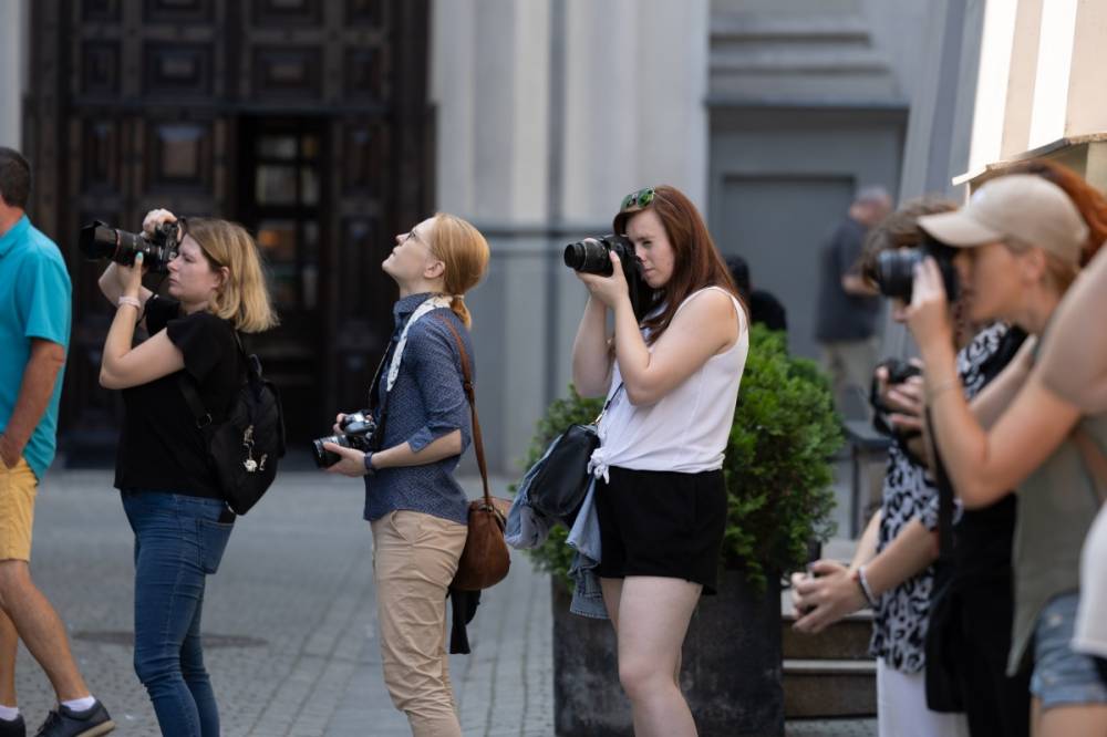 Grupa kobiet stojących na ulicy, każda z nich trzyma aparat fotograficzny i koncentruje się na robieniu zdjęć. Kobiety są różnie ubrane, niektóre mają na sobie letnie stroje, a inne bardziej casualowe. W tle widać fragmenty budynków oraz nieco rozmyte sylwetki przechodniów. Każda z fotografek wydaje się być skupiona na swoim celu, różne kąty ustawienia aparatów sugerują, że każda z nich fotografuje coś innego.