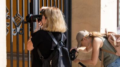 Dwie kobiety skupione na robieniu zdjęć w miejskim otoczeniu. Kobieta po lewej stronie, z jasnymi włosami, robi zdjęcie przez metalową kratę, celując obiektyw aparatu w stronę wnętrza lub detalu znajdującego się za kratą. Ma na sobie czarną koszulkę i nosi plecak. Kobieta po prawej stronie, w beżowej czapce i z jasnobrązowym plecakiem, pochyla się, aby uchwycić coś nisko przy ziemi swoim aparatem. Zdjęcie ukazuje moment w trakcie fotograficznej eksploracji detali architektonicznych.