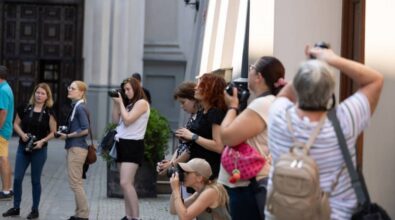 Grupa osób stojących na ulicy przed budynkiem, skupionych na robieniu zdjęć. Większość z nich to kobiety, trzymające w rękach aparaty fotograficzne. Są one w różnym wieku i mają różnorodne fryzury oraz stroje. Niektóre z nich przyjęły różne pozycje, aby uchwycić odpowiednie kadry – jedna z kobiet kuca, podczas gdy inne stoją. W tle widoczne są fragmenty architektury budynku oraz donice z roślinami.