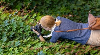 Kobieta w średnim wieku, która leży na brzuchu wśród gęstej zieleni roślin, trzymając aparat fotograficzny. Skupiona na robieniu zdjęcia, ma na sobie niebieską koszulę z drobnym wzorem i nosi torbę na ramię z brązowej skóry. Jej włosy są związane w kucyk.