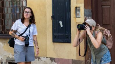 Dwie kobiety stoją przed zniszczonym budynkiem o żółtej fasadzie. Obie trzymają aparaty fotograficzne. Kobieta po lewej stronie ma na sobie biało-czarną bluzkę w paski, szare szorty i okulary. Jej ręce oraz nogi są pokryte tatuażami, a aparat wisi na pasku na jej szyi. Kobieta po prawej stronie, ubrana w zieloną koszulę bez rękawów, dżinsowe szorty i beżową czapkę z daszkiem, jest w pozie fotografowania, trzymając aparat przy twarzy. Na plecach ma różowy plecak. Obie wyglądają na skupione na wykonywanych zdjęciach. W tle widoczna jest obdrapana ściana budynku z kratą w oknie i ciemną skrzynką na ścianie.