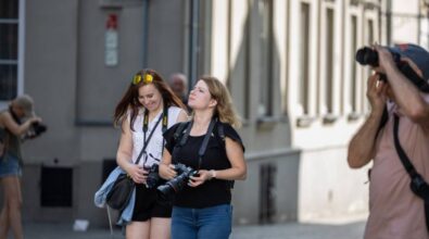 Dwie młode kobiety idą ulicą z aparatami fotograficznymi. Jedna z nich patrzy w górę z lekkim uśmiechem, trzymając aparat, podczas gdy druga również uśmiechnięta przegląda coś na ekranie swojego aparatu. W tle po lewej stronie widać mężczyznę w beżowej czapce, który także robi zdjęcia, pochylając się nad czymś przy ziemi. Cała scena rozgrywa się w miejskim otoczeniu, wśród budynków o jasnych elewacjach.