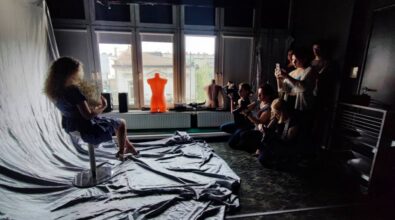 Zdjęcie przedstawia scenę z sesji fotograficznej w profesjonalnym studio. Na pierwszym planie widać lampy oświetleniowe, wentylator oraz statywy. W tle znajduje się duże okno, przez które wpada naturalne światło. Na środku siedzi młoda kobieta w niebieskiej sukience, trzymająca w dłoniach bukiet suchych kwiatów. Jest ona ustawiona na tle rozciągniętej białej tkaniny, tworzącej prostą scenografię. Po prawej stronie grupka osób robi zdjęcia.