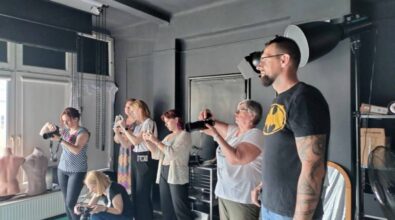 Zdjęcie przedstawia grupę ludzi w studio fotograficznym, skoncentrowanych na robieniu zdjęć. Widać kilka kobiet trzymających aparaty i smartfony, fotografujących obiekt znajdujący się poza kadrem. Pomieszczenie ma ciemne ściany, a na wyposażeniu widać profesjonalne lampy oświetleniowe, wentylator oraz manekiny ustawione przy oknie. Panuje atmosfera skupienia i kreatywnej pracy.